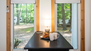 A room with a table set for breakfast, a lamp, and a view of a wooded area with trees and a path.