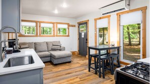 A modern kitchen with a grey sofa, wooden flooring, and large windows with views of trees.
