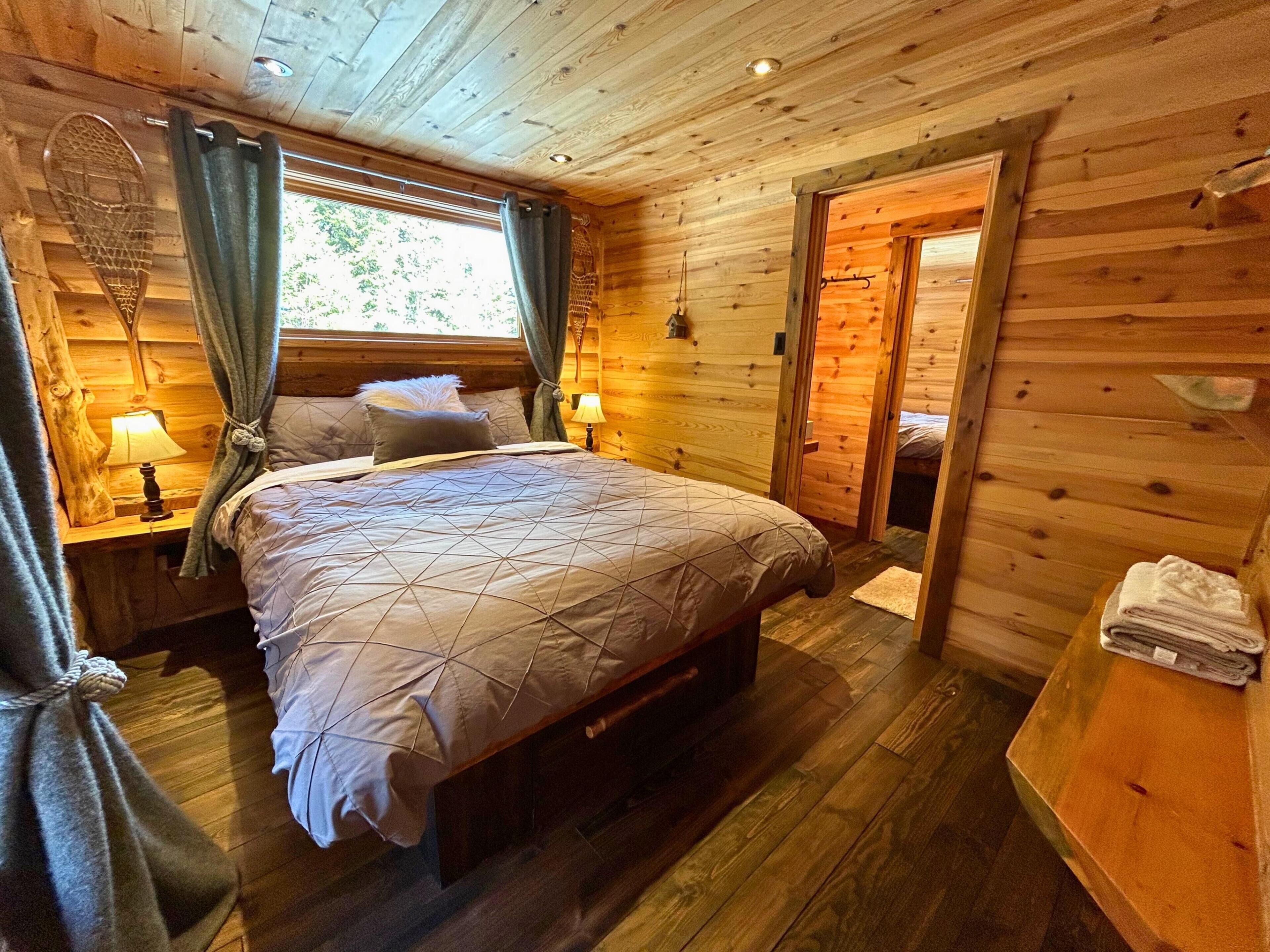 Quarto de cabana de madeira com cama, mesas de cabeceira e uma porta que dá para outro cômodo.