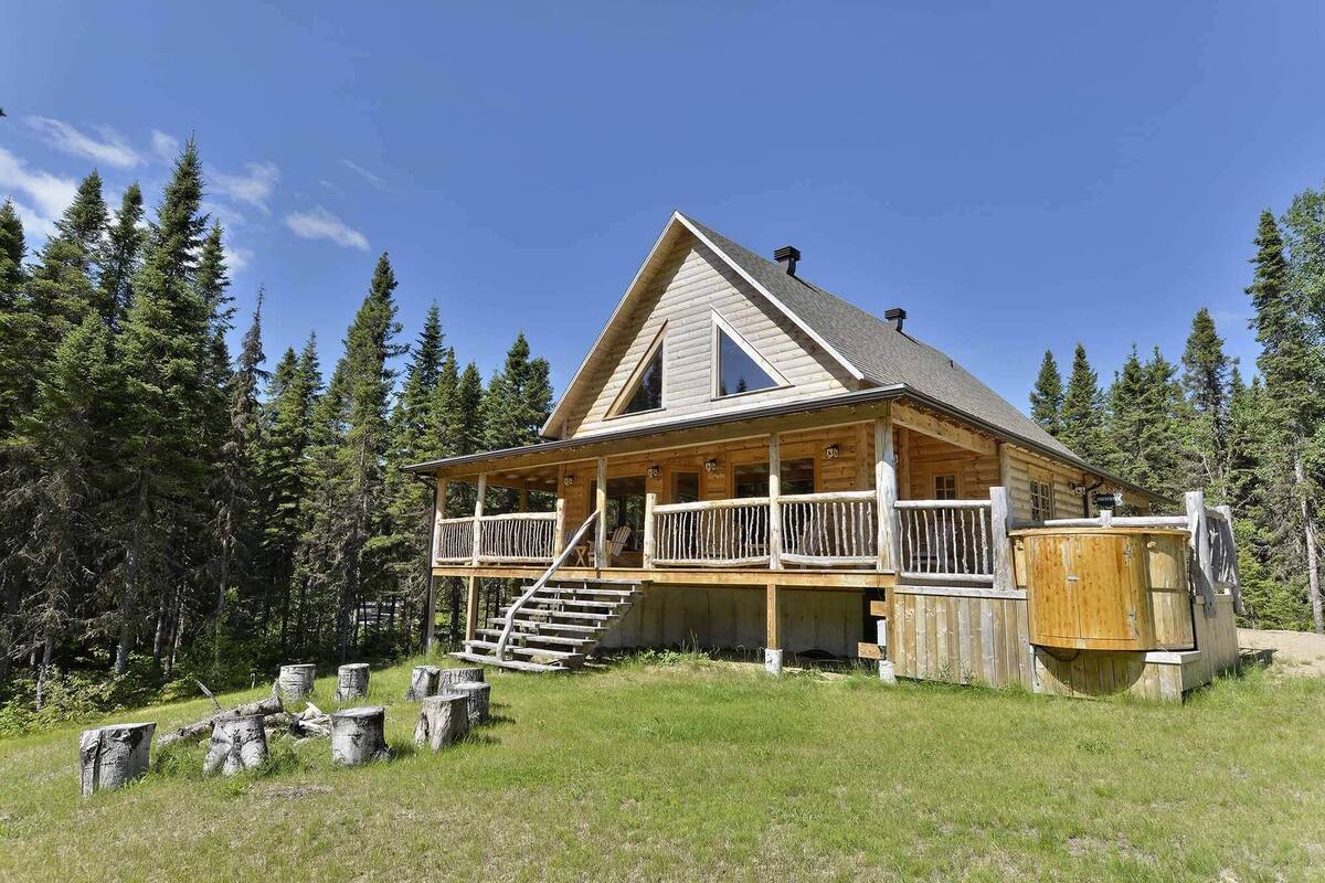Une cabane en rondins avec un porche, un escalier menant à l’entrée et un grand tonneau en bois à proximité.