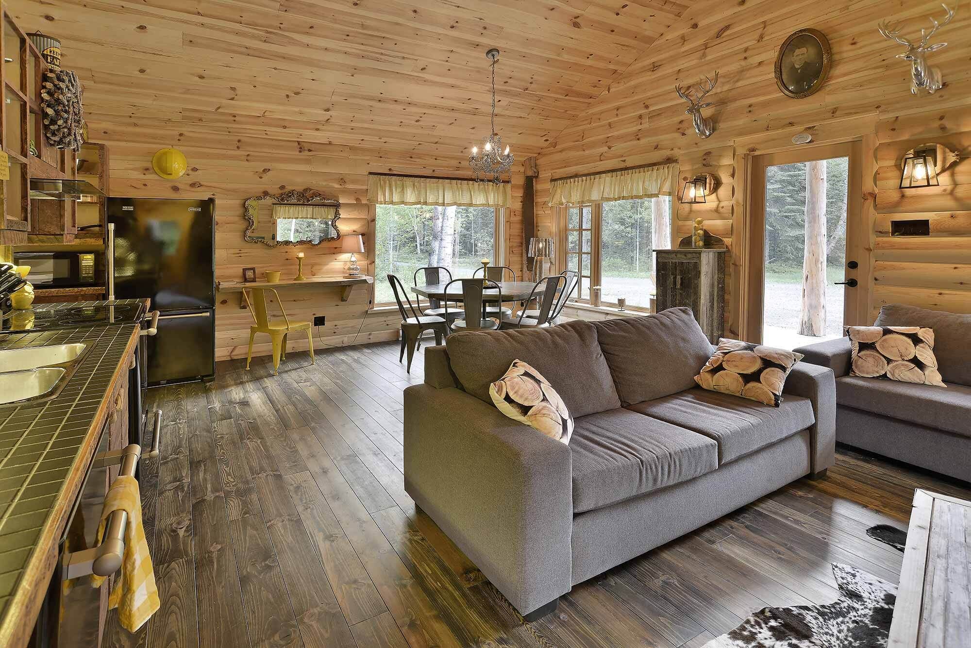 Interior acolhedor de uma cabana, com espaço de jantar, um sofá com almofadas decorativas e uma cozinha com frigorífico e piso de madeira.