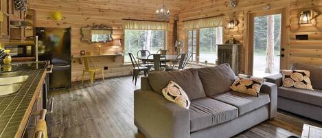 Interior acolhedor de uma cabana, com espaço de jantar, um sofá com almofadas decorativas e uma cozinha com frigorífico e piso de madeira.