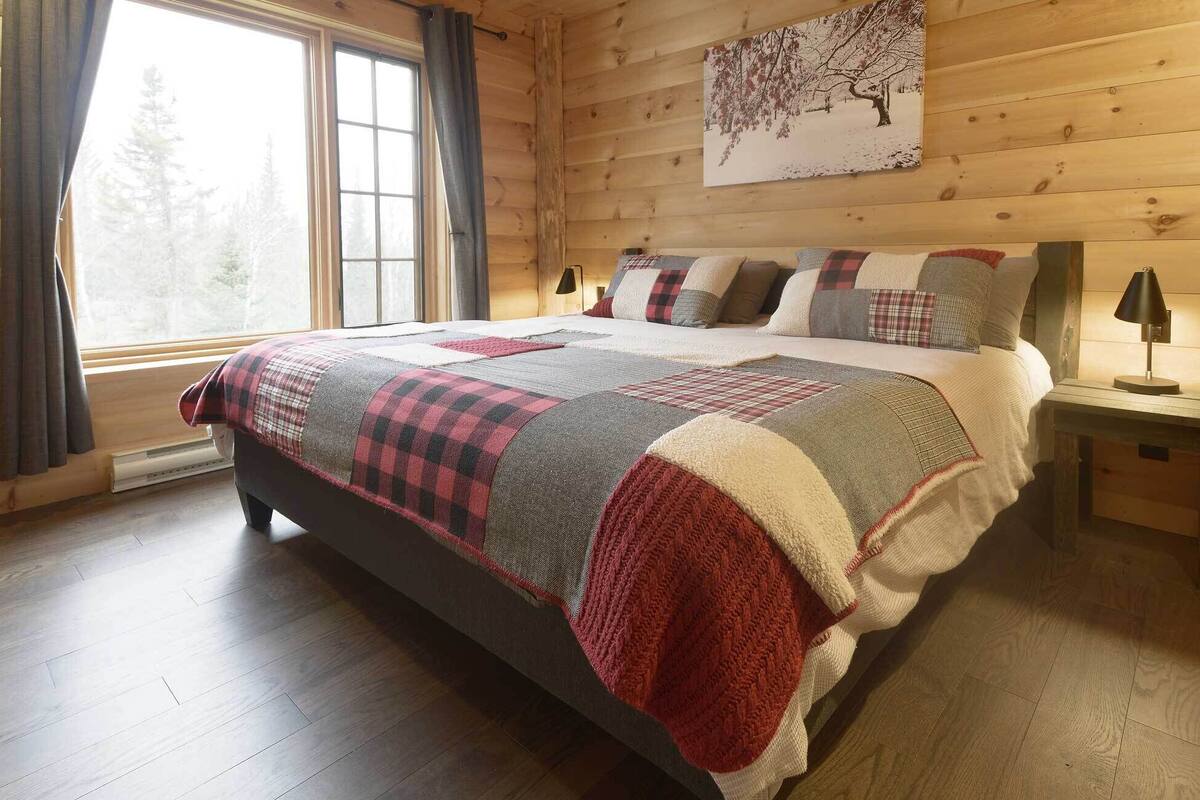 Une chambre dans une cabane en bois, avec un grand lit, une fenêtre avec des rideaux, un tableau au mur et une table de chevet avec une lampe.