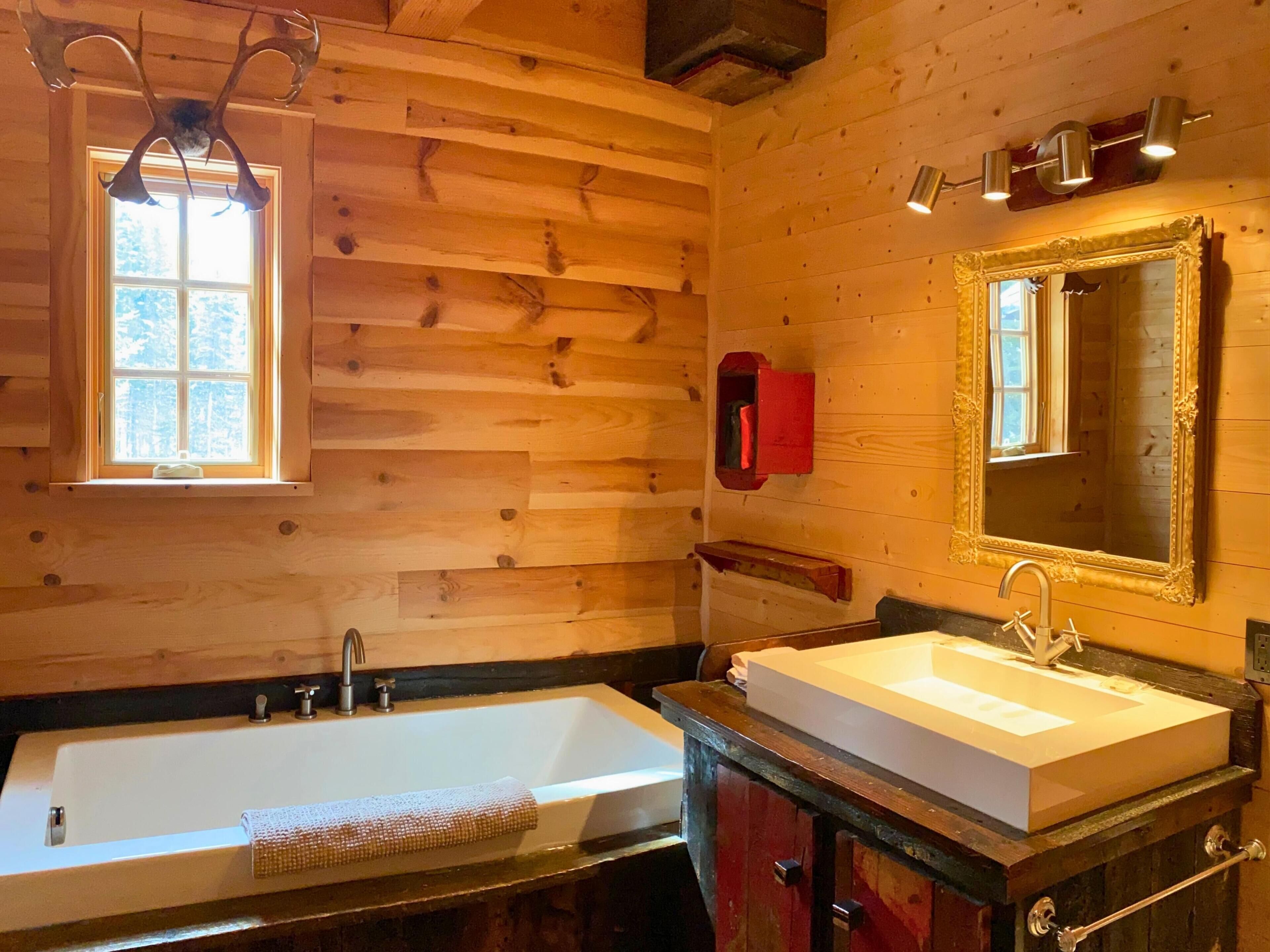 A wooden cabin bathroom with a bathtub, a sink, a mirror, and a window.