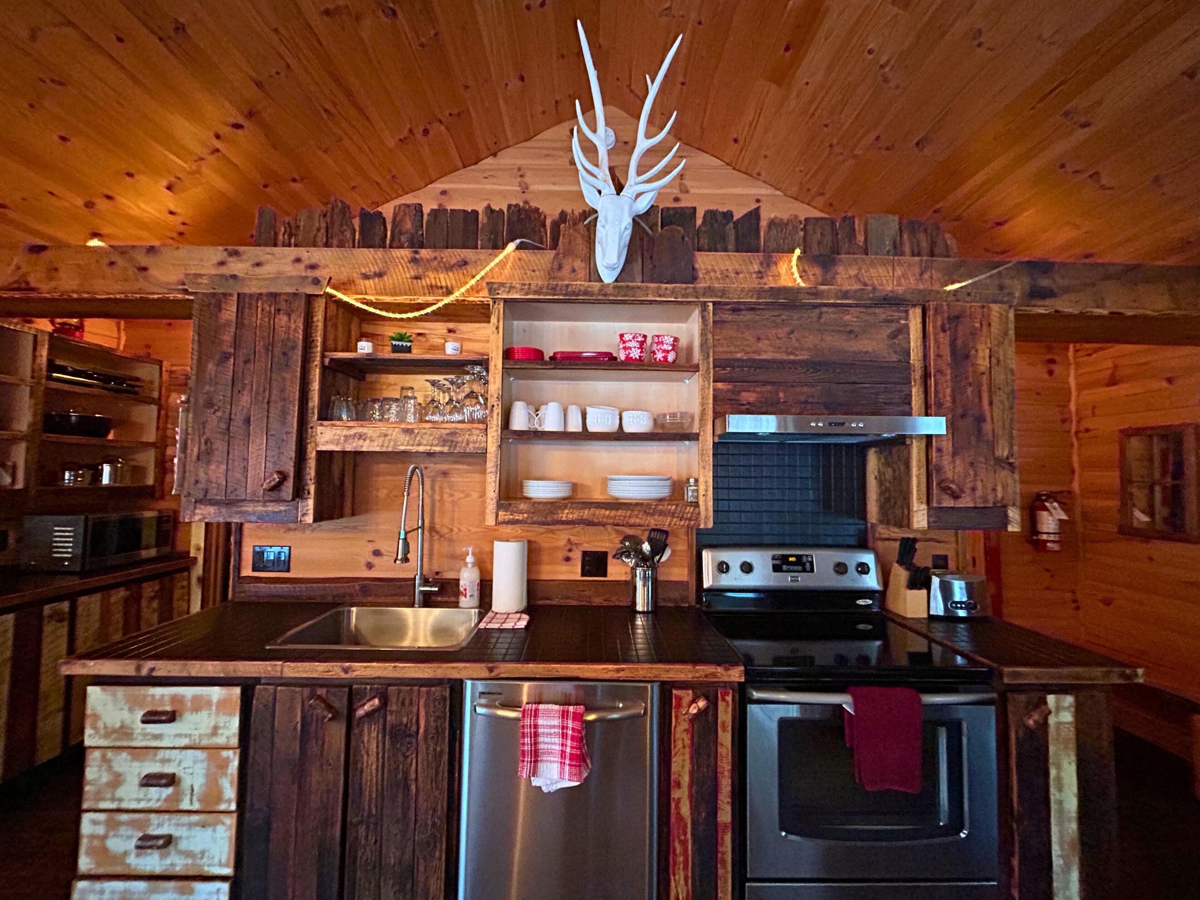 A rustic kitchen with wooden cabinetry, a stainless steel refrigerator, a stove, and a wooden ceiling with a deer head decoration.