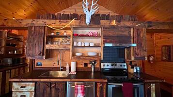 A rustic kitchen with wooden cabinetry, a stainless steel refrigerator, a stove, and a wooden ceiling with a deer head decoration.