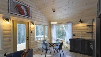 Interior de una cabaña de troncos con una mesa de comedor, sillas, un sofá y una zona de cocina.