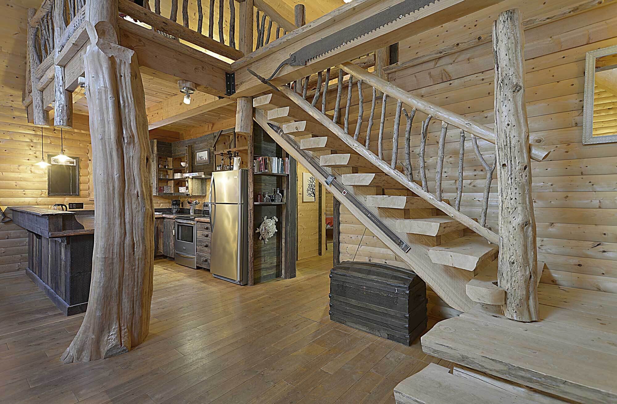 A rustic cabin interior with a wooden staircase, a kitchen area, and a dining space.