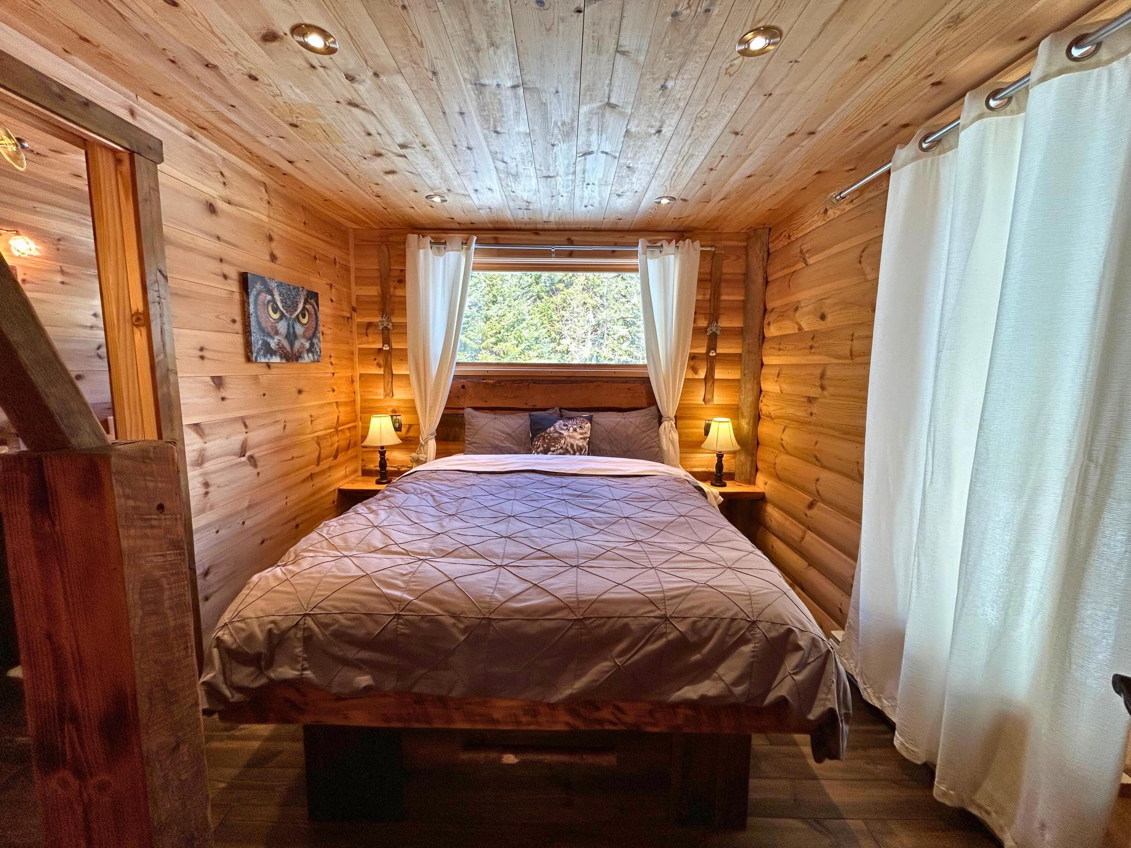 Une chambre à coucher dans un chalet en bois, avec un lit, deux lampes de chevet, une fenêtre avec des rideaux et un tableau accroché au mur.
