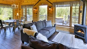 A cozy cabin interior with a fireplace, wooden furniture, and large windows offering a view of the forest. - Les Chalets de Môh  (La Malbaie)