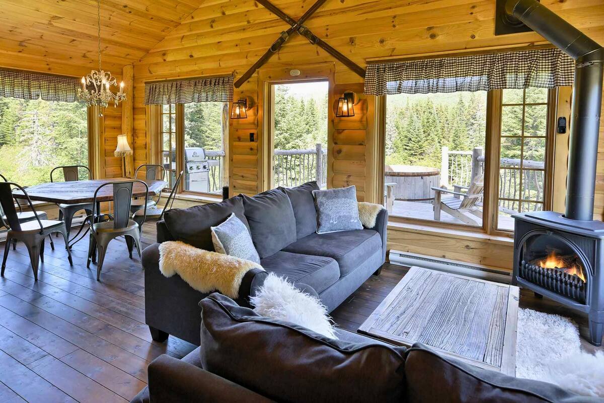 L’intérieur chaleureux d’un chalet avec une cheminée, des meubles en bois et de grandes fenêtres donnant sur la forêt.