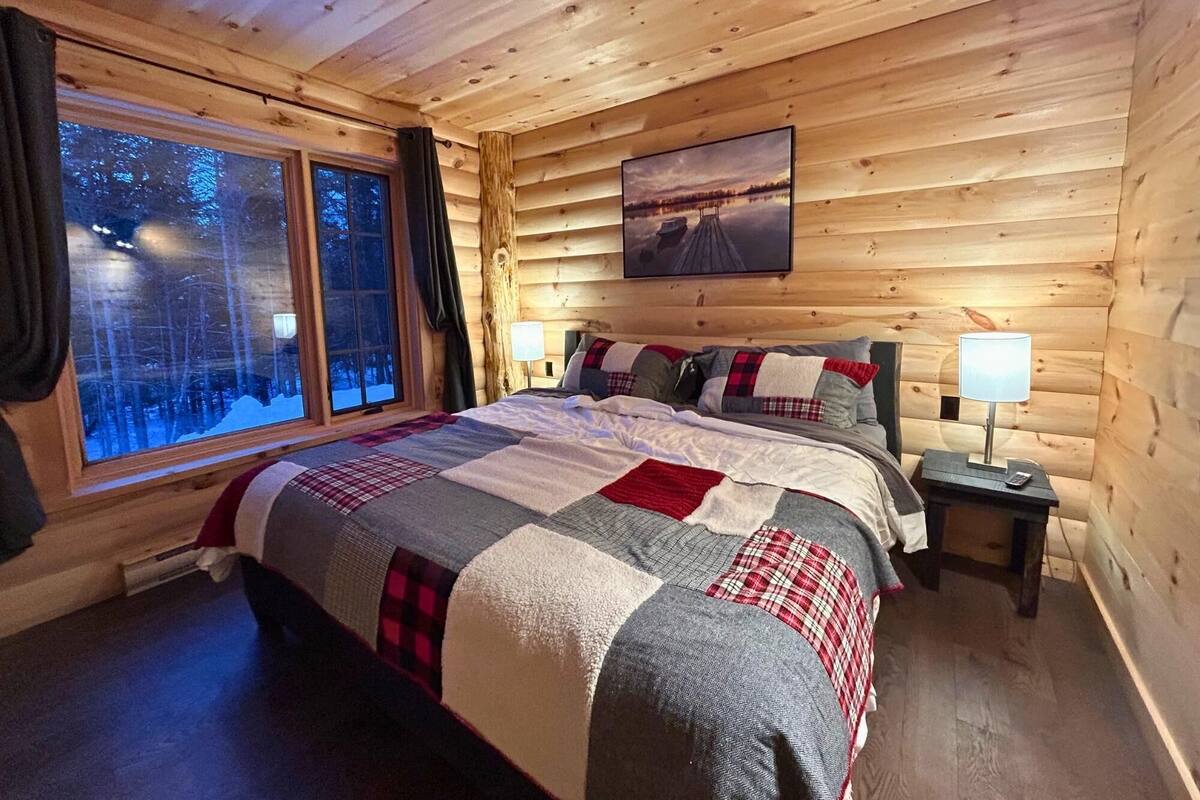 Une chambre confortable dans un chalet en rondins, avec un grand lit, une table de chevet, une fenêtre donnant sur les arbres et un tableau accroché au mur.