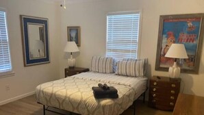 Een slaapkamer met een bed, twee nachtkastjes, een dressoir, een lamp, een ingelijst beeld en een raam met jaloezieën.