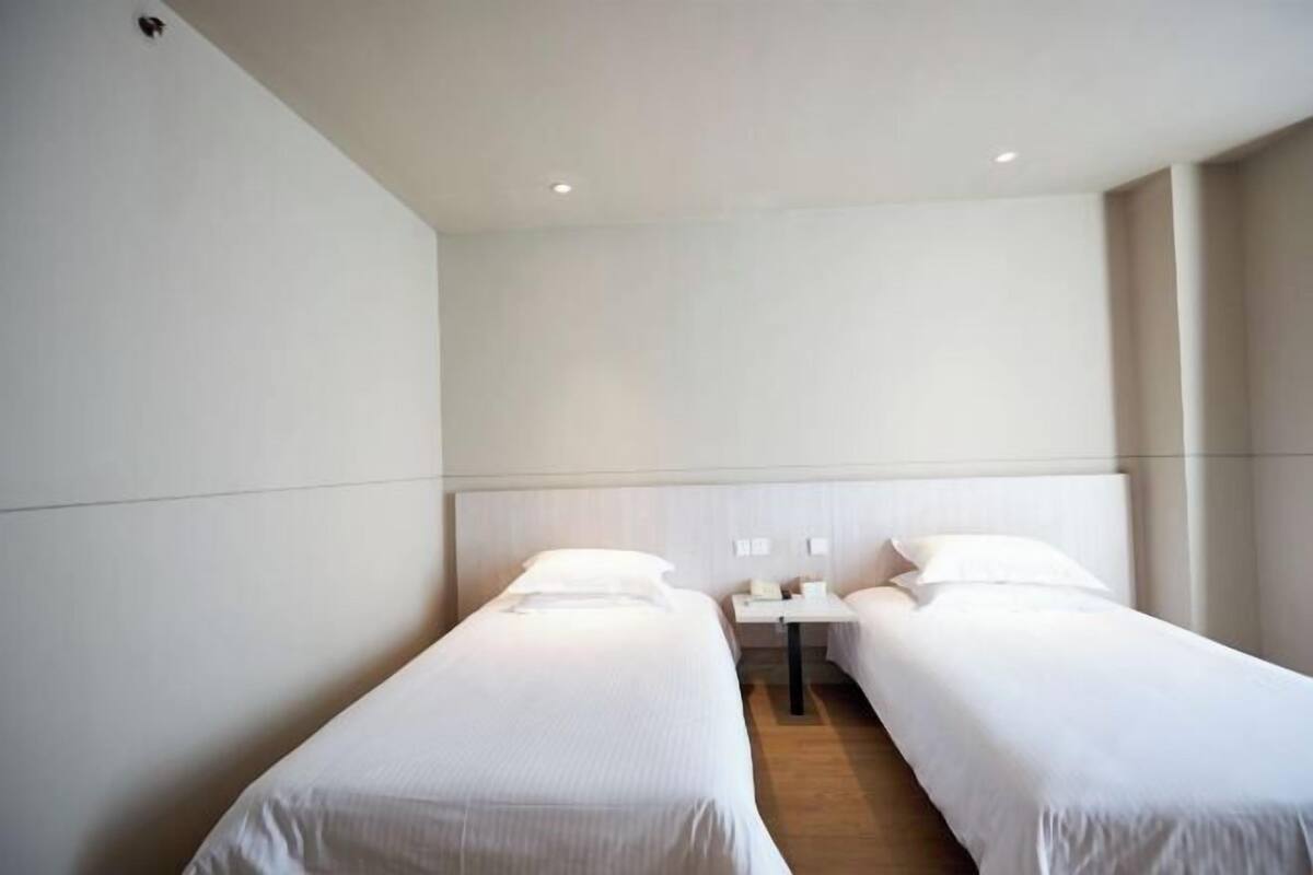 A hotel room with two single beds, a small bedside table with a lamp, and a plain white wall.