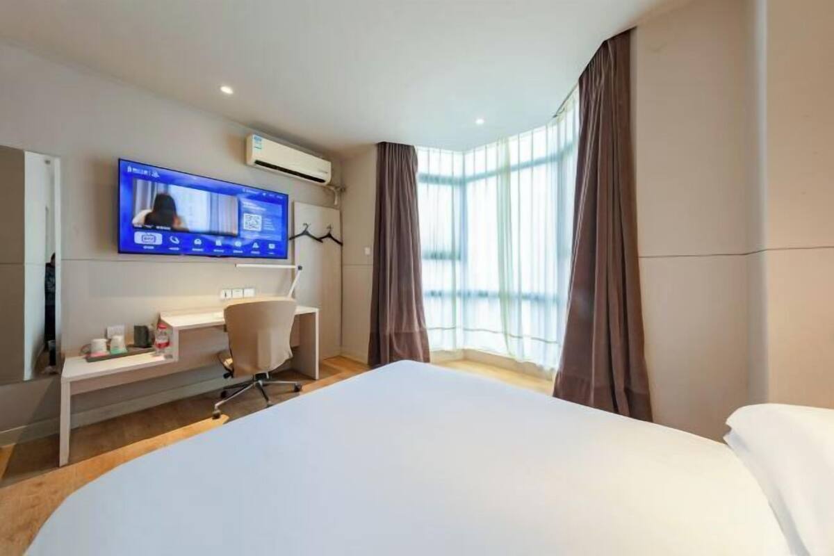 A hotel room with a bed, a desk, a chair, a TV mounted on the wall, and a window with curtains.