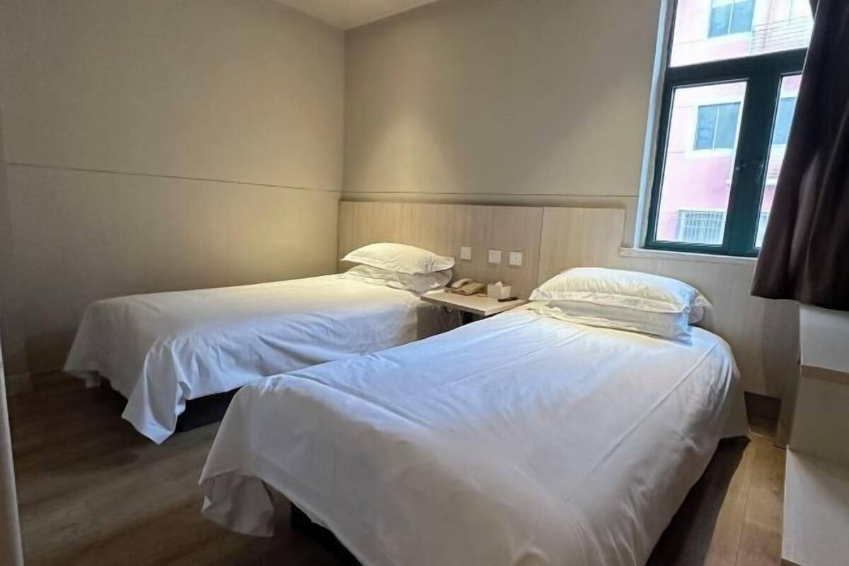 A hotel room with two single beds, a wooden nightstand, and a window with a view of buildings.