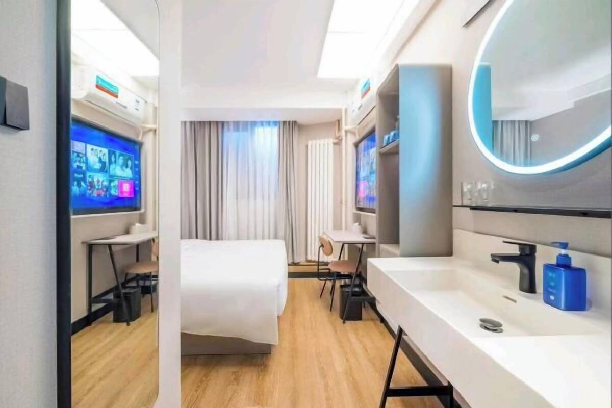 A modern hotel room with a bed, a desk, a chair, a large mirror, and a television screen displaying multiple channels.