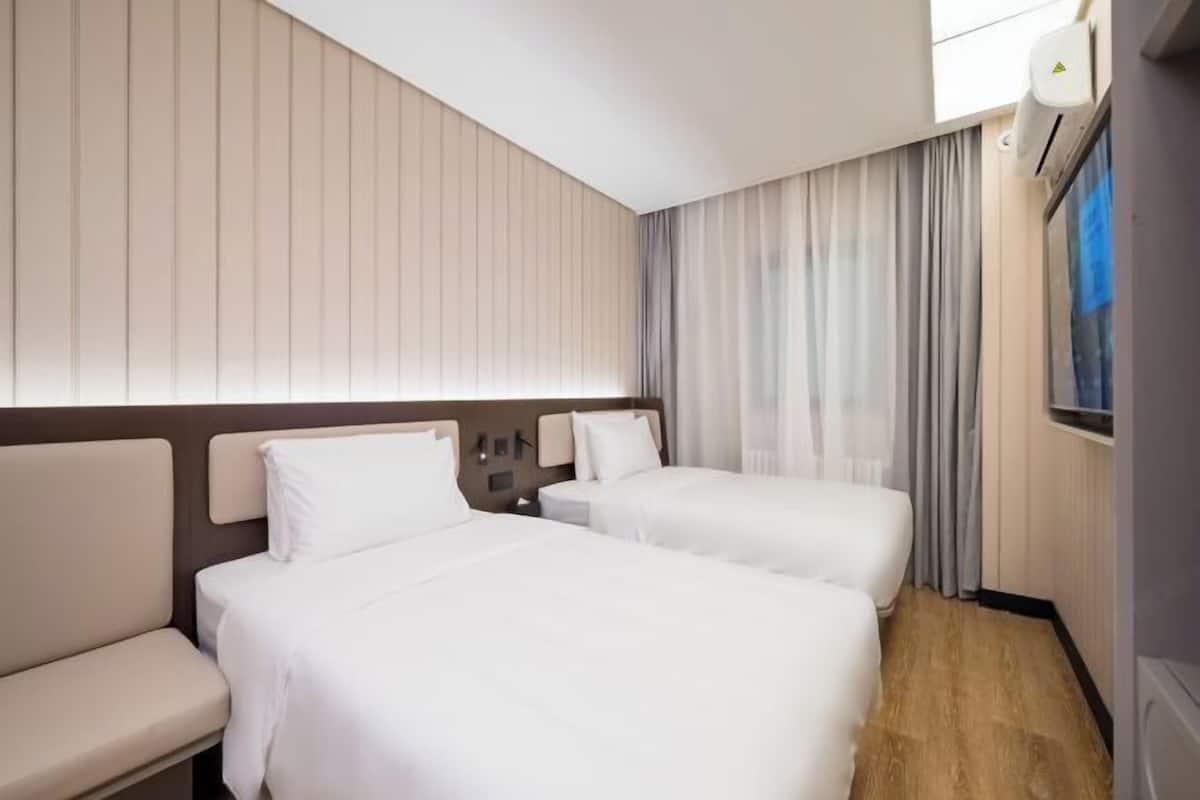 A hotel room with two beds, a sofa, a nightstand, and a wall-mounted air conditioning unit.