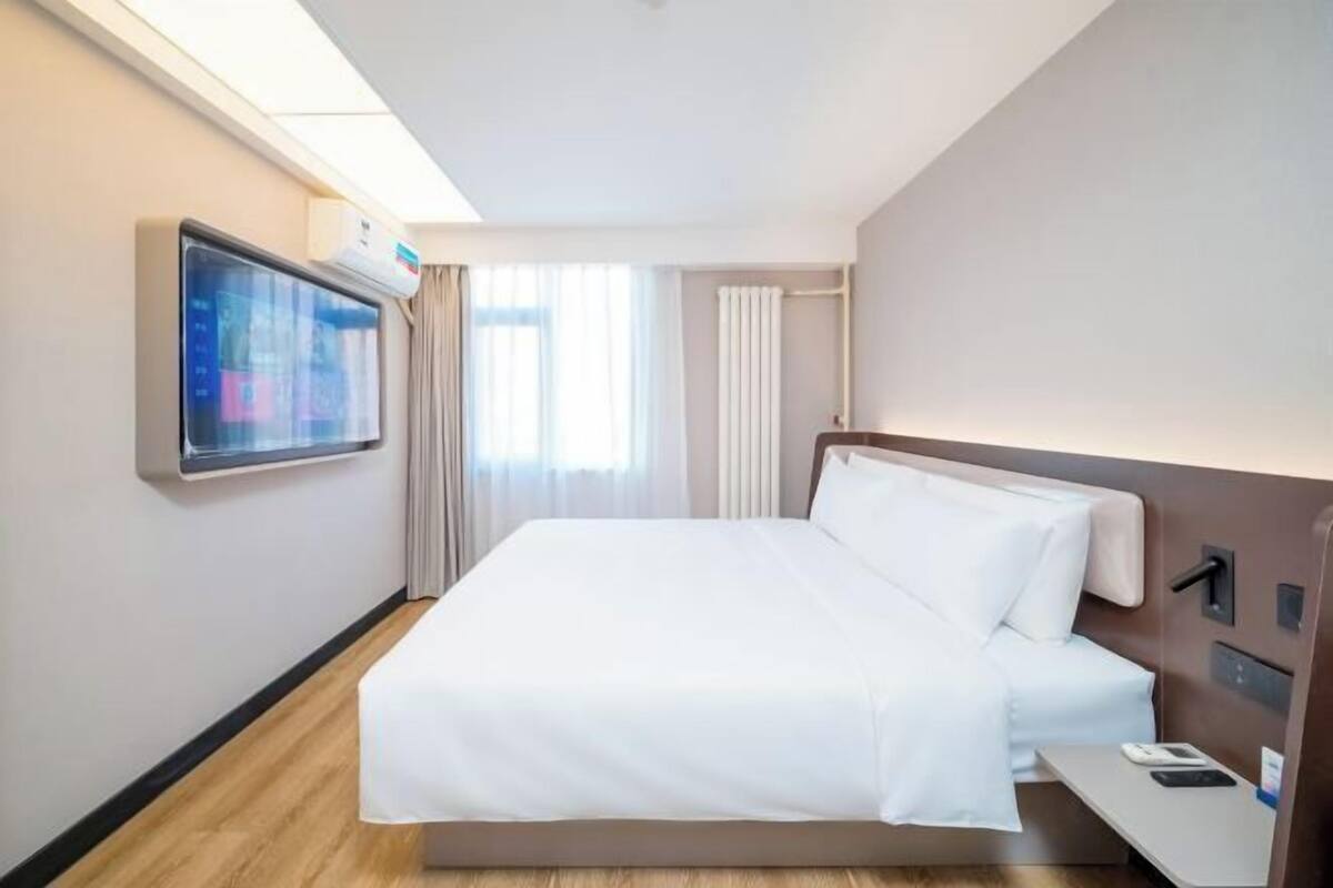 A hotel room with a bed, a TV mounted on the wall, a bedside table, and a window with curtains.
