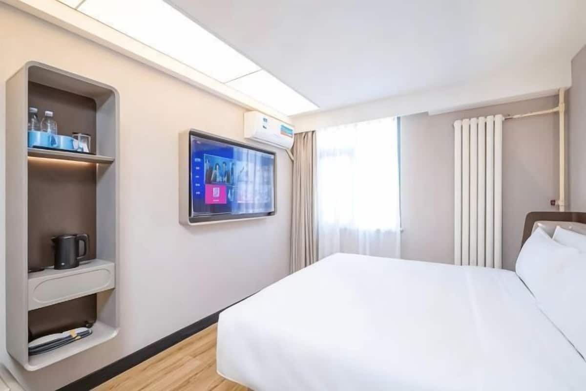 A hotel room with a bed, a TV mounted on the wall, a shelf with a kettle, and a window with curtains.