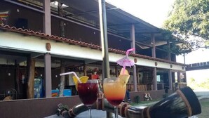 A bar area with two drinks on a table, a cocktail shaker, and a building with a tiled roof in the background. - Pousada Paraiso Azul (Anchieta)