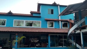 A two-story building with a blue exterior and a tiled roof. There are multiple windows, some with curtains, and a staircase leading to an upper level. - Pousada Paraiso Azul (Anchieta)