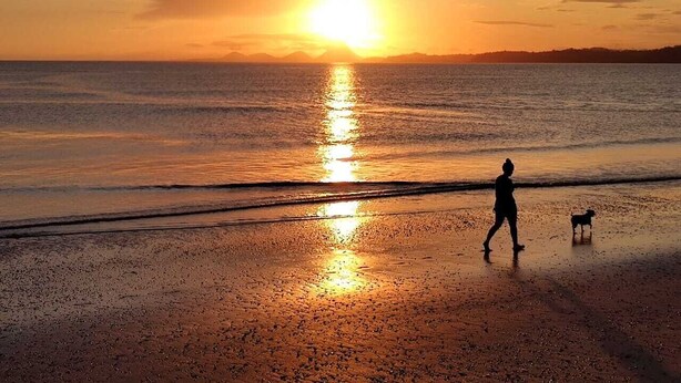 A beach scene with a sunset, a person walking a dog, and a calm sea. - Pousada Paraiso Azul (Anchieta)