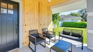 Un'area esterna moderna con posti a sedere, una porta nera, pareti in legno e vista su un prato verde e una casa sullo sfondo.