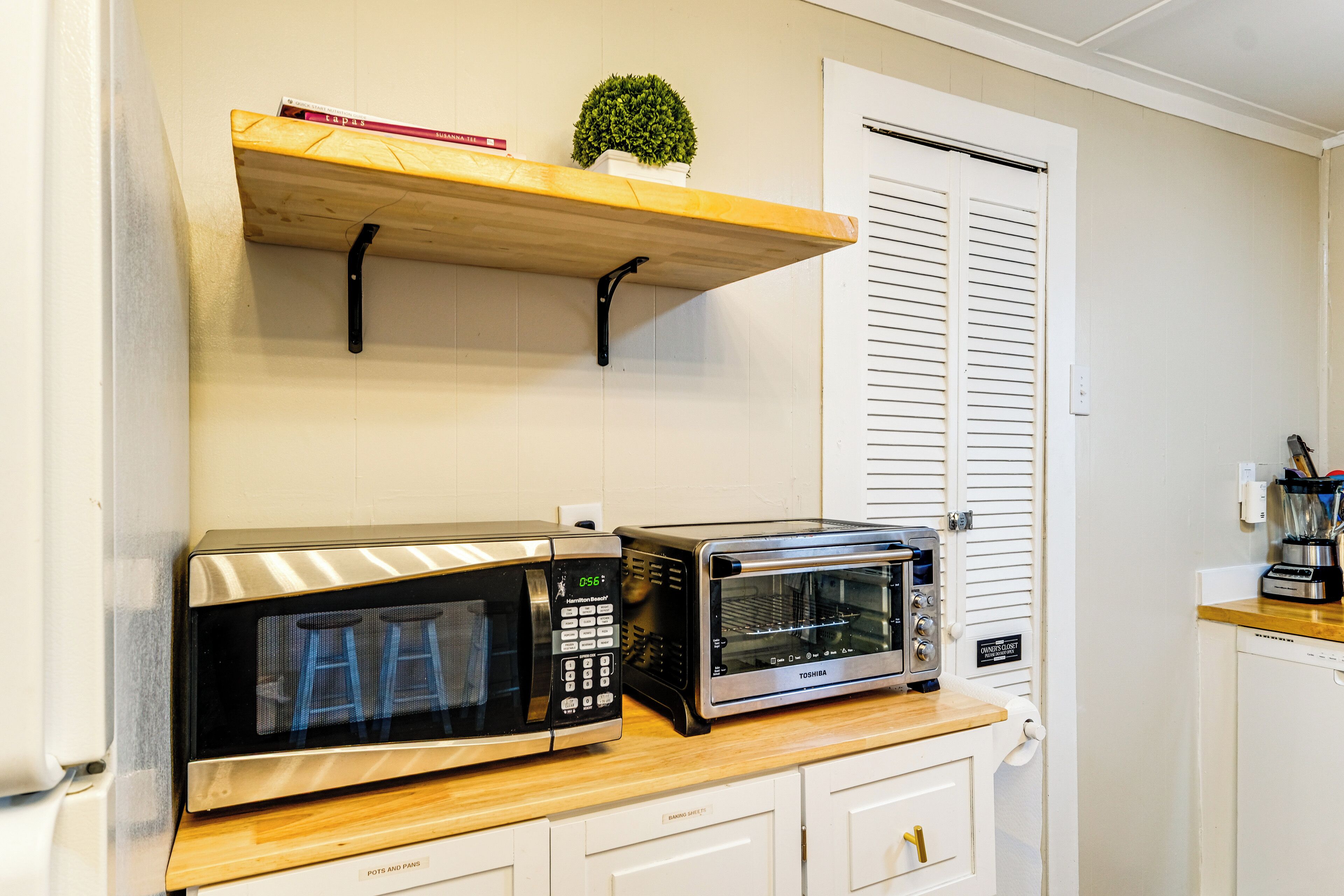 Une cuisine équipée d’un four à micro-ondes, d’un grille-pain et d’une étagère où sont posés des livres et une petite plante.