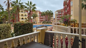 A balcony with a table and chairs overlooking a pool and hotel buildings.