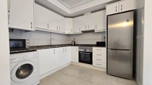 A modern kitchen with white cabinets, a stainless steel refrigerator, a built-in oven, a microwave, and a washing machine.