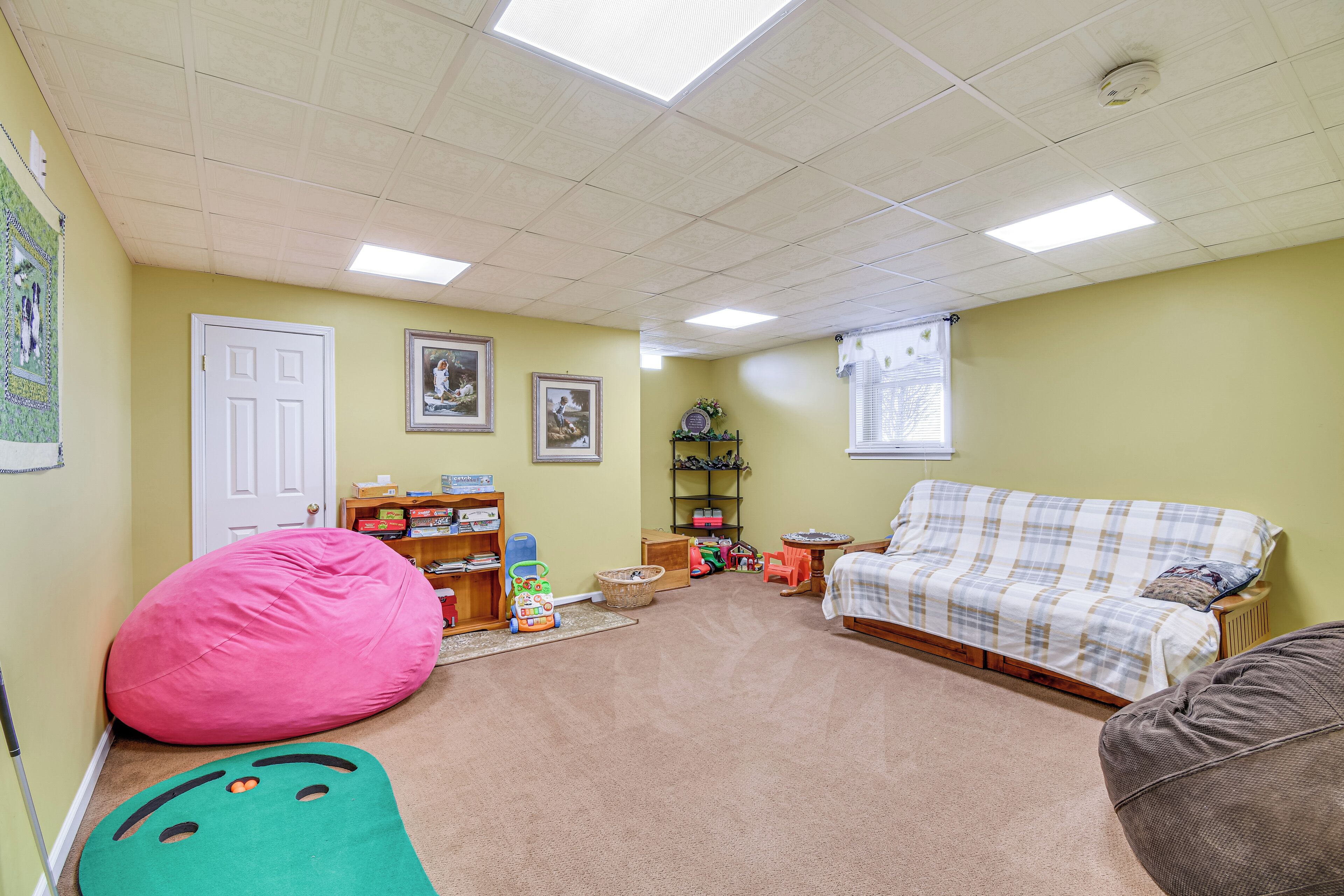 A room with a beige carpet, a pink bean bag, a green play mat, a plaid sofa, and a wooden bookshelf.