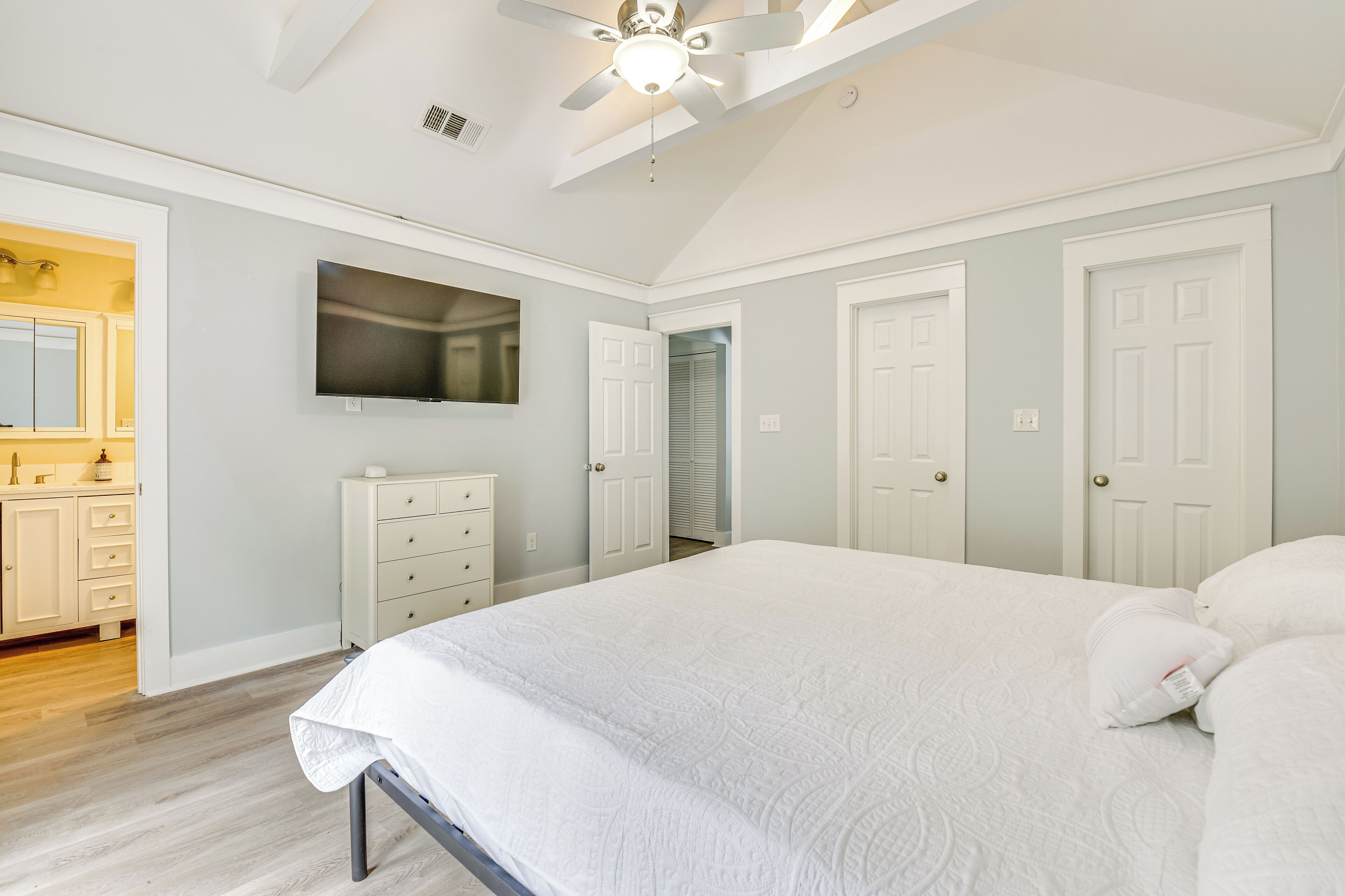 A bedroom with a bed, a ceiling fan, a TV mounted on the wall, and a door leading to a bathroom.