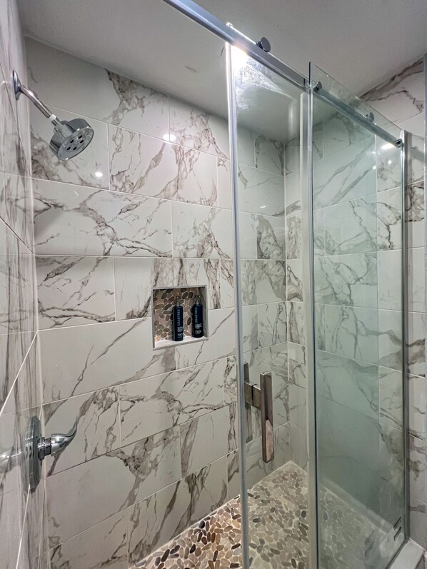 A modern bathroom with marble tiles, a glass shower enclosure, and a shelf with toiletries.