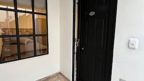 A room with a tiled floor, a black door with a number plate, and a window with blinds.