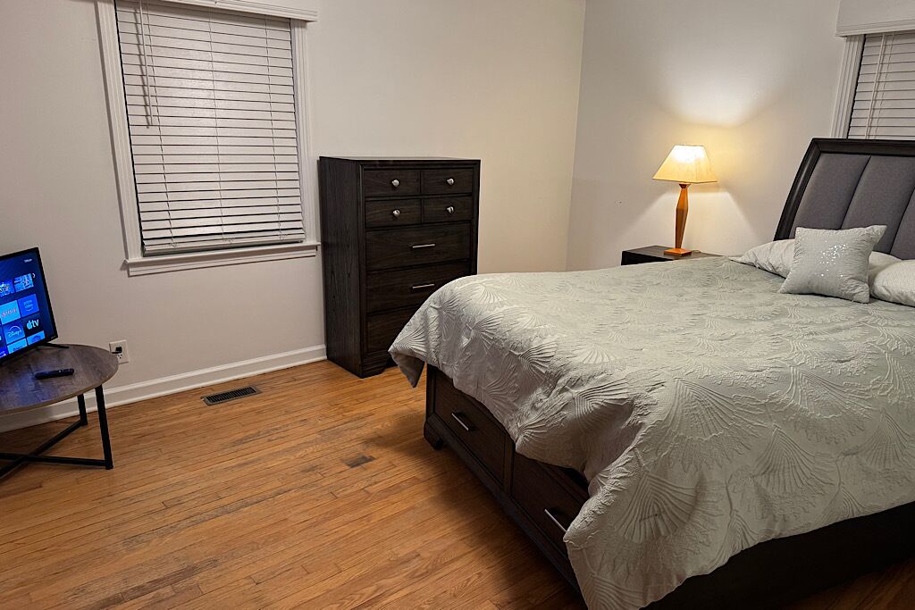 Een slaapkamer met een bed, een dressoir, een nachtkastje met een lamp en een televisie op een standaard.