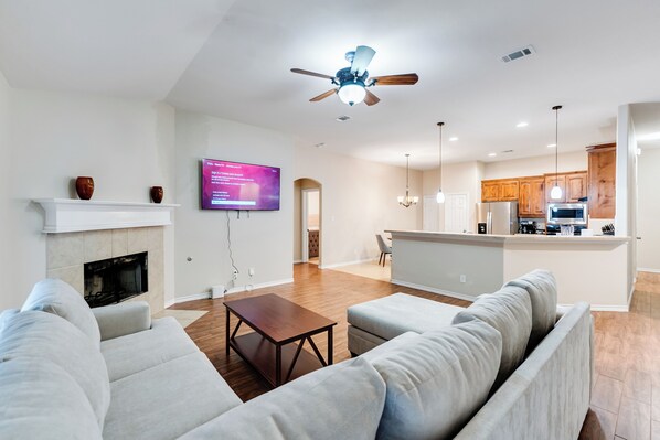A modern living room with a fireplace, a ceiling fan, and a kitchen in the background. - Relaxing Burleson Retreat · Pool Table & Space&5TV/3BA (Burleson)