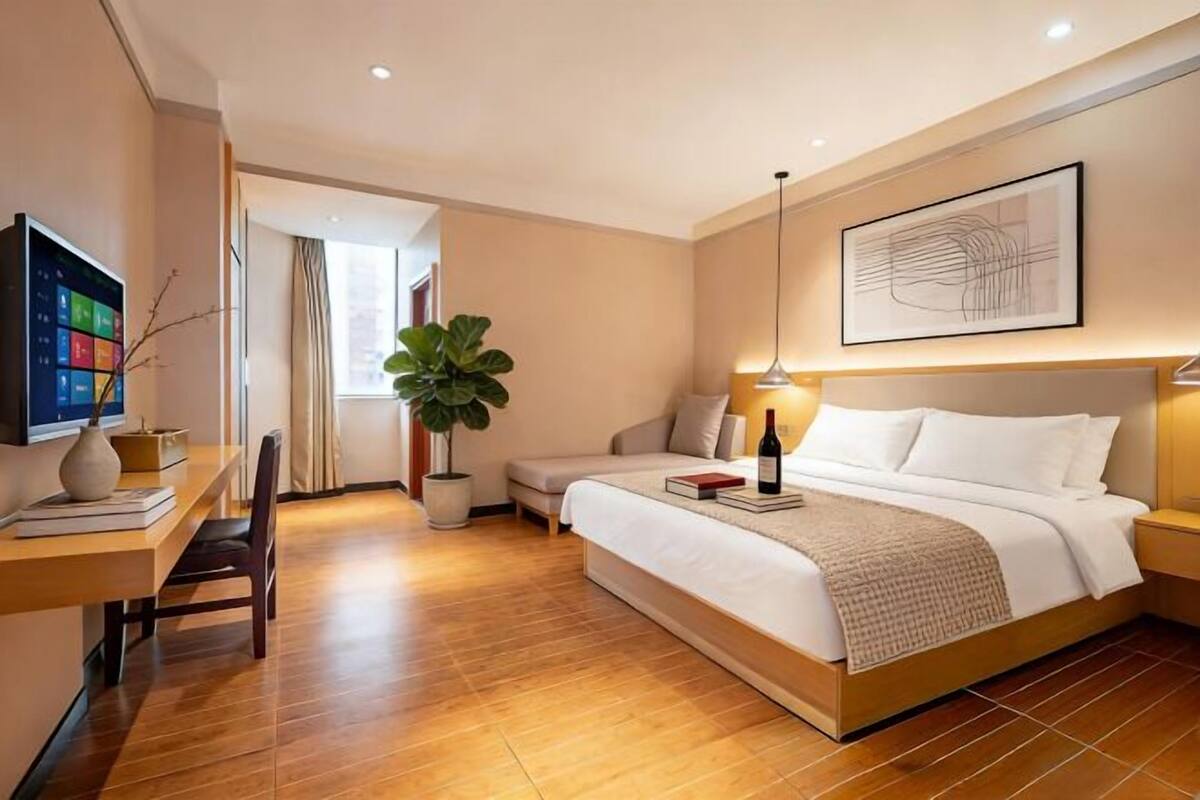 Quarto de hotel moderno com piso de madeira, cama com lençóis brancos, escrivaninha com computador, planta em vaso e uma pintura na parede.