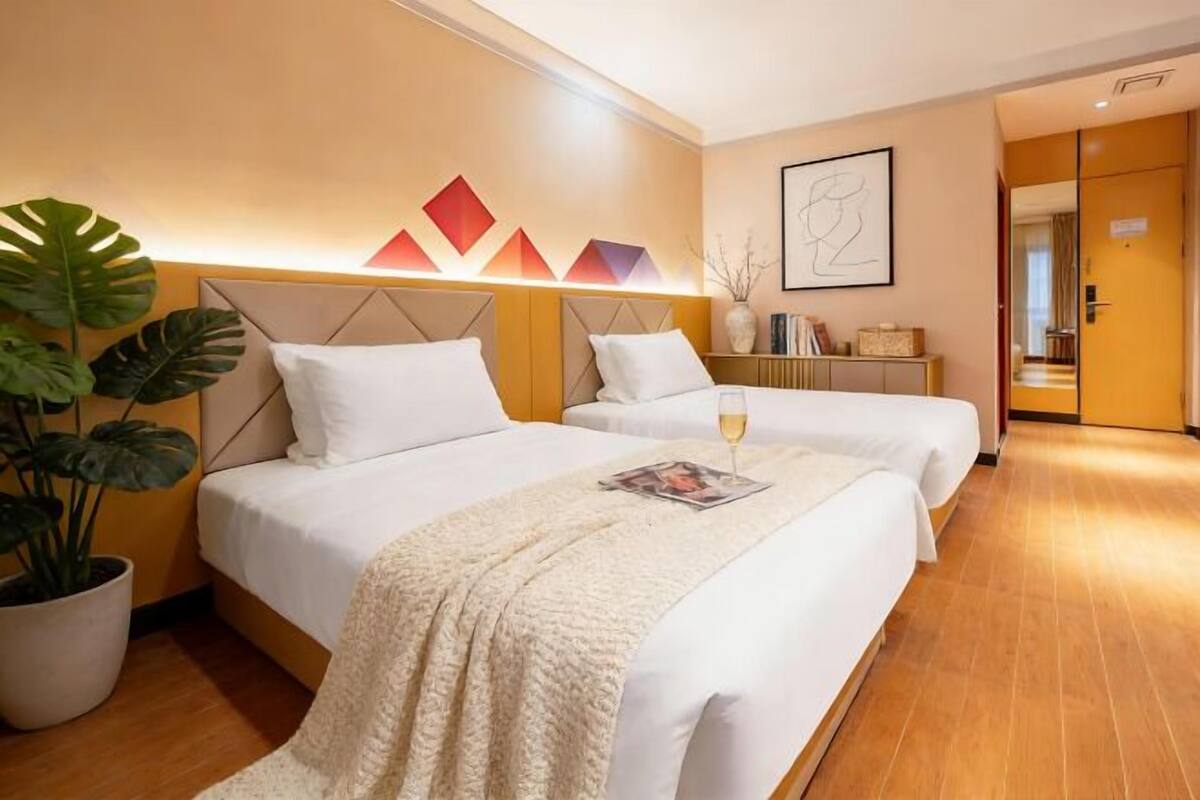 Quarto de hotel com duas camas, uma planta em vaso, um quadro emoldurado e uma mesinha de cabeceira.