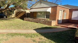 A single-story brick house with a front porch, a visible garage door, and a small front yard with grass and a dirt patch.