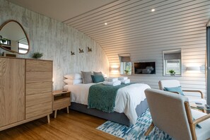 Un dormitorio moderno con un aparador de madera, una cama con sábanas blancas y una manta verde, una silla y un televisor montado en la pared.