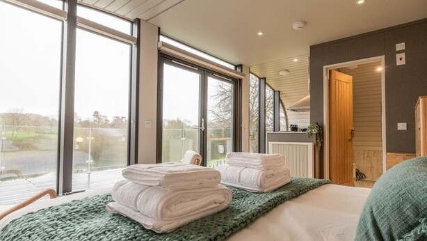 Una habitación de hotel moderna con ventanas amplias, una cama con ropa de cama verde y vista a un lago.