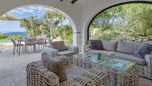 A covered outdoor seating area with wicker furniture and a glass table, overlooking a natural landscape with trees and a view of the mountains. - Mas Nou CostaBravaSi - Pool, AC, nature, views (Platja d'Aro)