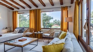 A cozy living room with a sofa, chairs, a coffee table, and a lamp. Large windows with curtains provide natural light and a view of greenery outside. - Mas Nou CostaBravaSi - Pool, AC, nature, views (Platja d'Aro)