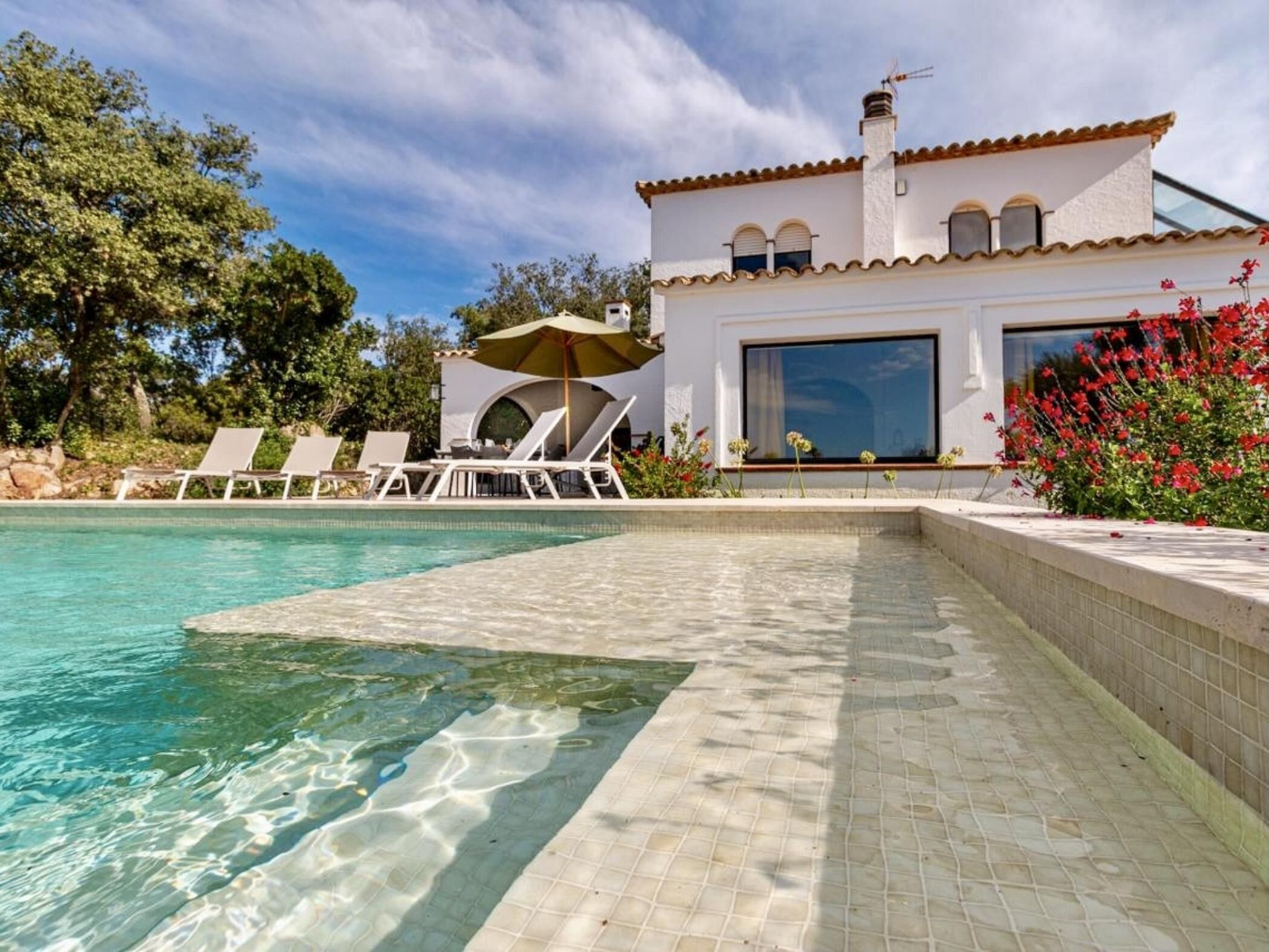 A poolside area with clear water, lounge chairs, and a white building with a tiled roof.
