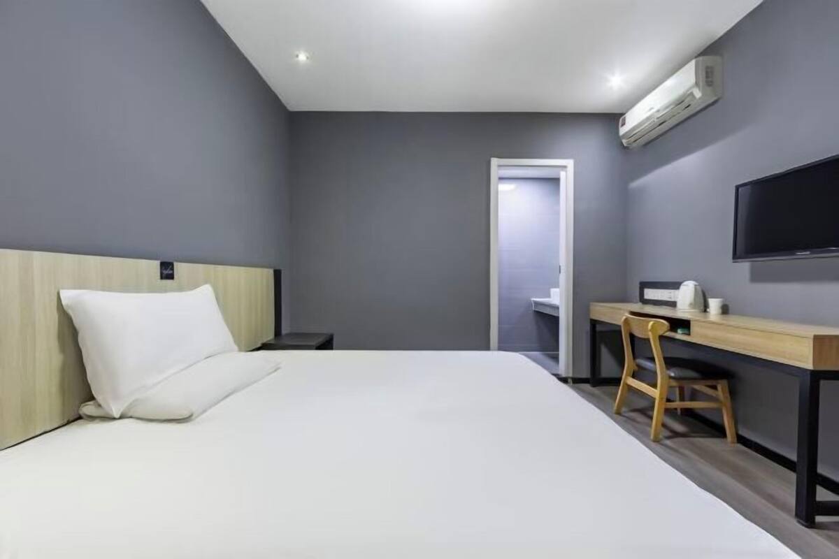 A hotel room with a bed, a desk, a chair, a TV, and an air conditioning unit.