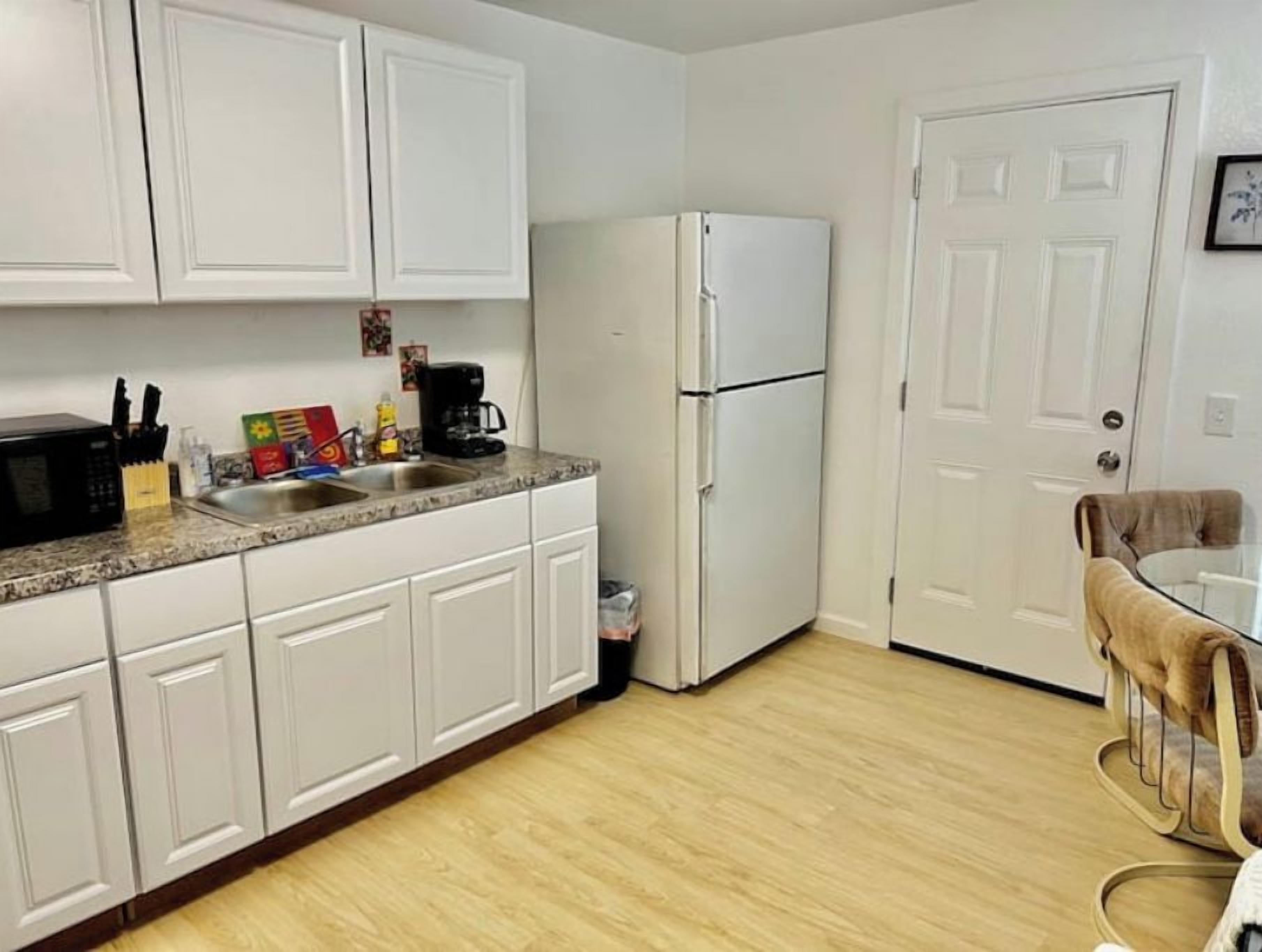 A kitchen with white cabinets, a refrigerator, a sink, and a dining area with a chair.
