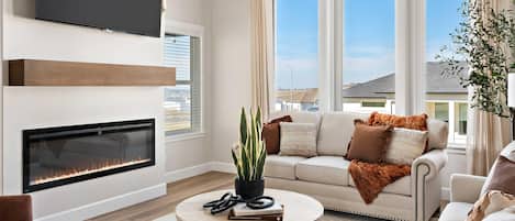 A modern living room with a fireplace, a flat-screen TV mounted on the wall, a large window with a view of the city, and a chandelier.