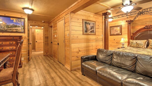 Interior de una cabaña de madera con sofá de cuero, paredes de madera, ventilador de techo y un cuadro enmarcado en la pared.