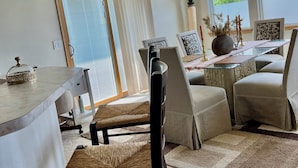 Ein Essbereich mit Tisch, Stühlen und einer Chandelüster. Ein Fenster mit Jalousien, eine Pflanze und dekorative Gegenstände stehen auf dem Tisch.